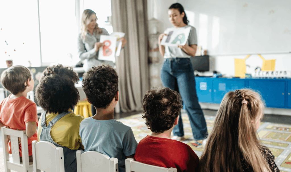 Crianças assistem aula com professoras mostrando letras, ressaltando a necessidade de um canal de denúncias na educação desde os primeiros anos escolares.