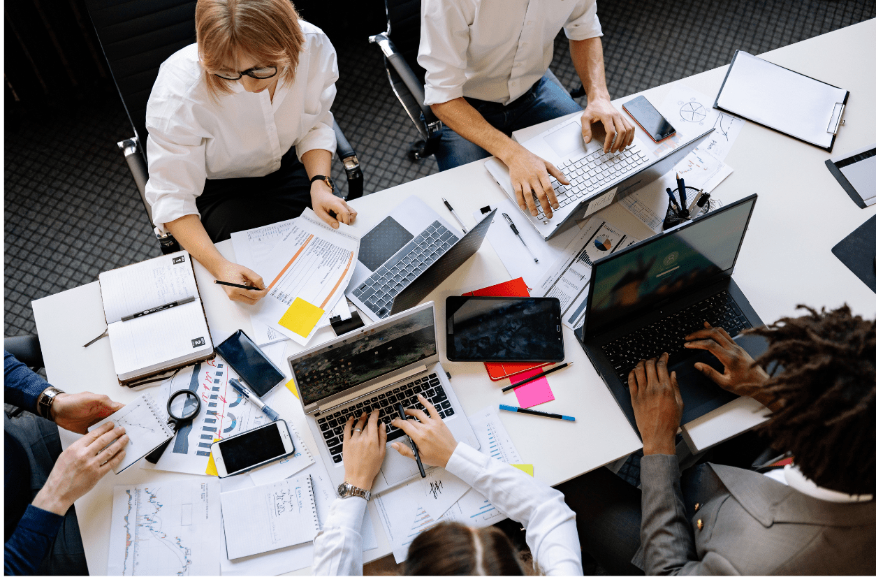 Pessoas reunidas em mesa de trabalho com computadores e documentos, simbolizando a implementação do canal de denúncias e Lei 14.457/2022 nas empresas.