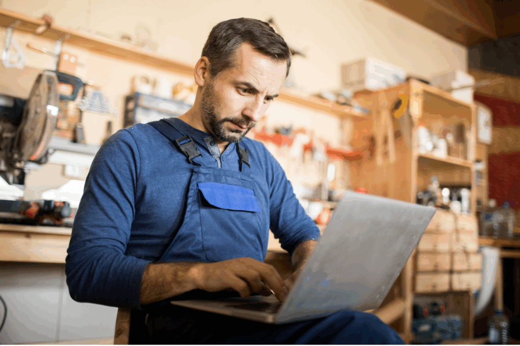 Homem em uma oficina de marcenaria trabalhando em um laptop. Ele está focado em seu trabalho e o ambiente ao redor sugere um espaço de trabalho criativo e técnico.