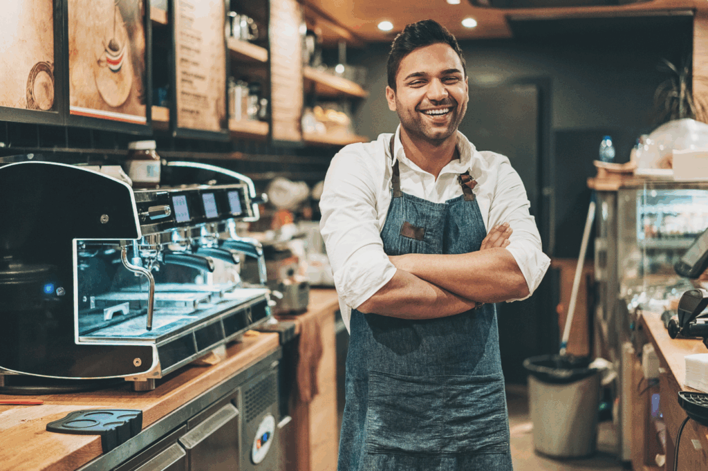 Barista feliz, usando um avental, sorrindo ao lado de uma máquina de café expresso em uma cafeteria. Canal de denúncias em PME pode ajudar a melhorar o ambiente de trabalho em estabelecimentos como este.