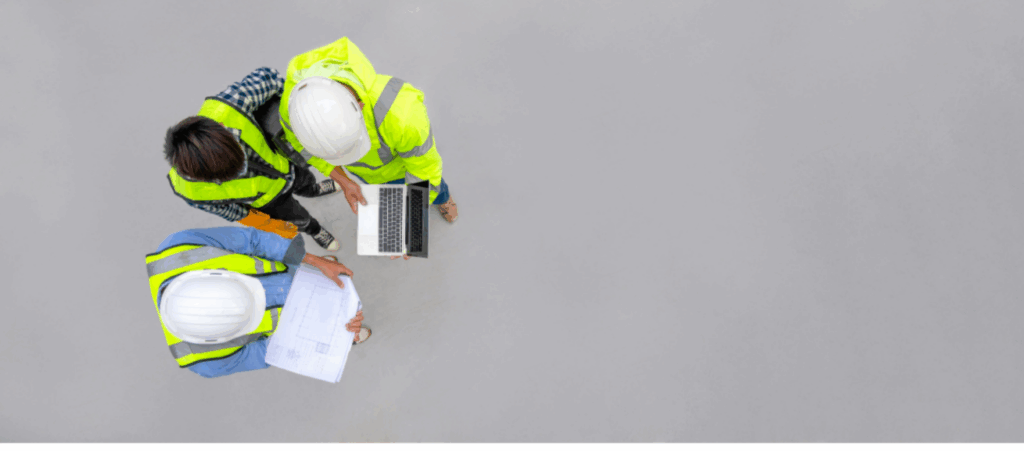 Três engenheiros com capacetes e coletes de segurança analisando planos de construção, com um laptop ao fundo. Canal de denúncias na construção civil é crucial para prevenir acidentes e garantir práticas seguras.