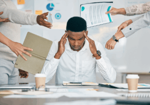 Profissional pressionado por múltiplas demandas simultâneas, com colegas entregando documentos e solicitações, cenário associado a canal de denúncias burnout assédio NR-1 e gestão de riscos ocupacionais.